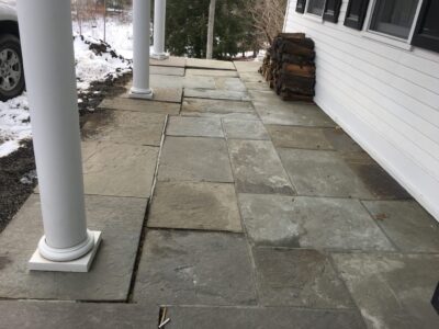a stone paver porch with uneven stones, indicating poor drainage, along with winter's freeze-thaw cycles, that caused pavers to become uneven. This not only detracts for the aesthetic appeal but also poses a serious tripping hazard.